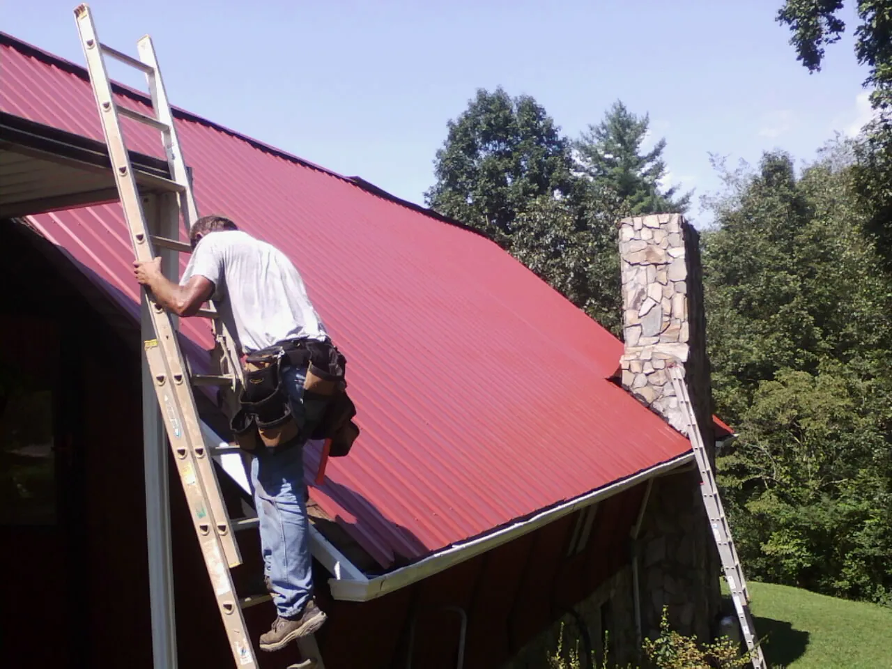 Roofer on a ladder beside a red metal roof and stone chimney — Soffit & Fascia Repair by Roofing Brothers in Bradenville